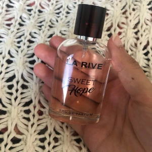 La Rive Sweet Hope Eau de Parfum - La Rive Sweet Hope Eau de Parfum med ljusrosa vätska i en genomskinlig glasflaska. Flaskan har rundade former och svart plastkork med silverdetalj. Snygg och feminin design, perfekt för dig som gillar söta dofter.