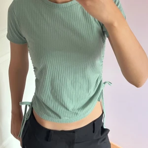 Ribbad grön topp med knyt  - Snygg ribbad topp i ljusgrön färg. Modellen är croppad med korta ärmar och har trendiga knytband i sidorna som ger en cool detalj. Perfekt för dig som gillar en enkel men ändå unik stil. Står storlek M men sitter mer som en xs/s