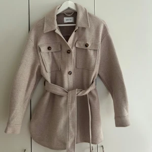 Beige kappa - Stilren beige overshirt med bälte i midjan och stora bröstfickor. Jackan har knappar framtill och klassisk krage, perfekt inför hösten.
