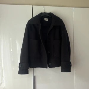 Svart kappa från Kappahl XS - Stilren svart jacka från Kappahl i ullblandning, storlek XS. Jackan har stora fickor framtill, bred krage och knappar både framtill och vid ärmslut. Perfekt för dig som gillar en klassisk look med modern touch.