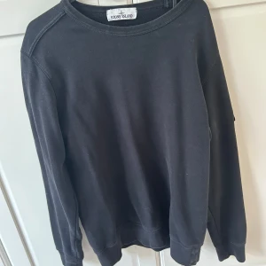 Svart sweatshirt från Stone Island - Svart långärmad sweatshirt från Stone Island med klassisk rund hals och ikonisk logotyp-patch på vänster ärm. Tröjan är i mjuk bomull och har en clean, stilren look som passar perfekt till jeans eller cargo. Snyggt avslut vid ärmslut och midja.