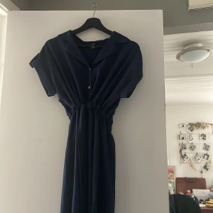 Marinblå långklänning med krage - Säljer en stilren marinblå långklänning från Monki med klassisk krage och korta ärmar. Klänningen har knappar framtill och ett snyggt rynkat midjeparti som ger en cool siluett. Perfekt för dig som gillar enkel men trendig design.