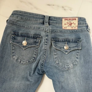 True Religion ljusblå bootcut jeans - Säljer ett par ikoniska ljusblå jeans från True Religion med låg midja och bootcut. Jeansen har snygga ficklock med silverfärgade knappar och klassisk logga bak. Tillverkade i mjukt denim med coola sömdetaljer och en avslappnad vibe.