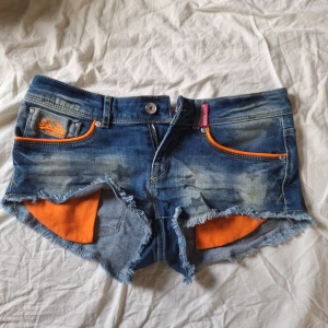 Superdry jeansshorts med stjärnor - Ascoola jeansshorts från Superdry med slitna kanter och coolt stjärnmönster på fickorna. De har orange detaljer vid fickorna och insidan, de är även väldigt lågmidjade. Perfekta för sommarens!