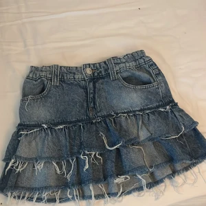 Jeans kjol 146-152 - Säljer en blå jeanskjol med coola volanger och råa fransar längst ner. Kjolen har klassiska fickor fram och bak, samt hällor för bälte. Perfekt för dig som gillar en avslappnad och trendig stil med lite edge.