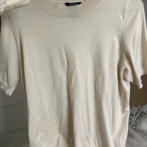 Beige stickad kortärmad tröja Lindex - En stilren beige stickad tröja från Lindex i storlek S. Tröjan har korta ärmar, rund halsringning och ribbade muddar vid hals och ärmslut. Perfekt för dig som gillar en clean och enkel look med mjukt material.