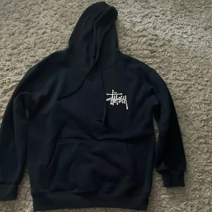 Mörkblå Stüssy hoodie med logga - Mörkblå hoodie från Stüssy med stor vit logga på ryggen och mindre logga på bröstet. Klassisk streetwear-stil med huva och känguruficka framtill. Mjuk och skön, perfekt för chill dagar. Långärmad och med ribbade muddar vid ärmslut och nederkant.