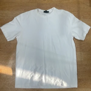 Vit t-shirt från SMOG, relaxed fit - Klassisk vit t-shirt från SMOG i relaxed fit. T-shirten har rund halsringning och korta ärmar, tillverkad i mjuk bomull. Enkel och clean design som funkar till allt, perfekt för dig som gillar en avslappnad stil.