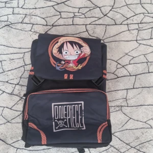 One Piece ryggsäck med Luffy-motiv - Cool ryggsäck med stort One Piece-tryck och Luffy på framsidan. Väskan är svart med orange detaljer och repmönster, har flera fack och vadderade axelremmar. Perfekt för animefans som vill sticka ut med sin stil.
