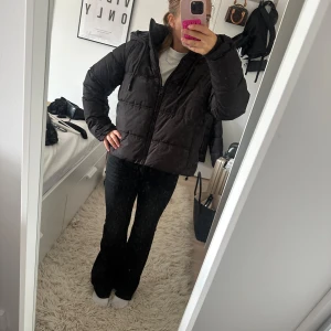 Svart pufferjacka med huva - Säljer en svart pufferjacka med huva och dragkedja framtill. Jackan har snörning i huvan och är quiltad med breda kanaler. Perfekt för kalla dagar och har en clean, enkel look som passar till det mesta.