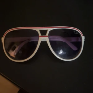 Vita Ray-Ban pilotglasögon med röd rand - Säljer ett par snygga Ray-Ban solglasögon i klassisk pilotmodell. Bågarna är vita med en röd rand längs ovansidan och mörka glas. Glasögonen har en cool retrostil och passar perfekt för dig som vill sticka ut med en unik look.