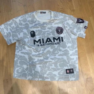Inter Miami x BAPE fotbollströja - Snygg Inter Miami fotbollströja i samarbete med A Bathing Ape. Tröjan är vit med ljusgrått camouflage-mönster, korta ärmar och tryckta loggor på bröstet och ärmen. Perfekt för dig som gillar streetwear och fotboll.
