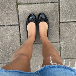 Svarta ballerinaskor med lacktå - Snygga svarta ballerinaskor med glansig lacktå och söt rosettdetalj framtill. Skorna har ett diskret rutigt mönster och är perfekta för en stilren och trendig look. Passar till både kjol och jeans.