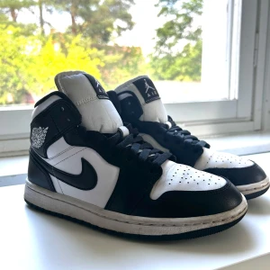 Nike Air Jordan 1 svartvita sneakers - Svartvita Nike Air Jordan 1 sneakers med klassisk hög siluett, svart swoosh och vita paneler. Skorna har snörning, perforerad tå och ikoniska Jordan-loggor på sidan och plös. Tillverkade i skinn och syntet, perfekt för dig som gillar streetstyle och basketvibe.
