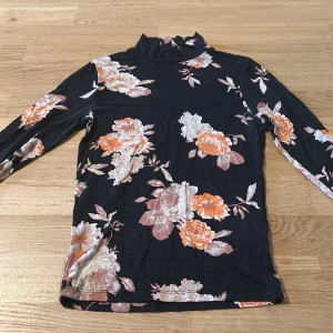Blommig polotröja från Gina Tricot - Supersnygg svart polotröja från Gina Tricot med stora blommor i orange, beige och vitt. Tröjan har lång ärm och smal passform, perfekt att styla med jeans eller kjol. Mjukt och stretchigt material som sitter skönt hela dagen.