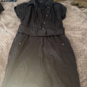 Svart långklänning från Hugo Boss - Stilren svart långklänning från Hugo Boss med klassisk krage, knappar framtill och bredt bälte i midjan. Klänningen har korta ärmar och fickor med knappdetaljer. Perfekt för dig som gillar en tidlös och elegant look. Hör av vid frågor❤️pris går att diskutera 