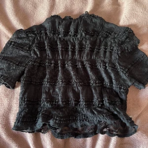 Svart spetsig croppad blus med volanger - Säljer en svart croppad blus med korta ärmar och hög hals. Blusen är helt i spets med flera lager volanger och transparenta partier