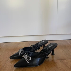 Svarta pumps med strassrosett från Pieces - Superfina svarta klackskor från PIECES i storlek 41! Köptes för 649kr.   De har en spetsig tå, kitten heel och är dekorerade med en glittrig strassrosett framtill. Använda endast vid ett tillfälle, så de är i mycket fint skick! Säljes pga fel storlek.  