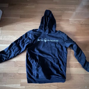 Svart Sail Racing hoodie - Svart hoodie från Sail Racing med vit logga på bröstet. Klassisk känguruficka framtill, huva med dragsko och mjukt material. Tillverkad i 78% bomull och 22% polyester. Perfekt för dig som gillar en clean och sportig stil.