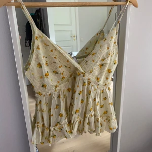 Blommig volangtop med spetsdetaljer - Supersöt blommig linnetopp med tunna axelband, volanger och spetsdetaljer. Toppen är ljusgul med små gula blommor och har smock i ryggen för extra komfort. Perfekt för varma dagar och ger en romantisk vibe.