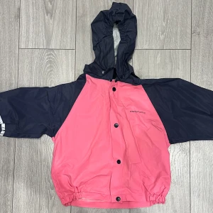 Rosa och marinblå regnjacka Prosports för barn - Snygg tvåfärgad regnjacka från Prosports i rosa och marinblått. Jackan har huva, tryckknappar framtill och elastisk nederkant. Perfekt för regniga dagar och enkel att slänga på sig över andra kläder. Oversized känsla med långa ärmar storleken har vi ingen aning om men det skulle nog passa 4-5 åringar. 