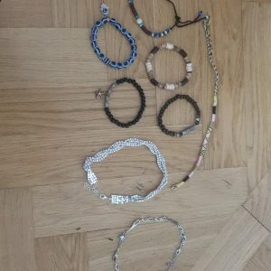 Mix av armband och halsband i boho-stil - Säljer ett set med flera armband och ett halsband. Armbanden är i olika färger och material: blått pärlarmband med ögon och hamsa, träarmband i brunt och beige, svart pärlarmband med sjöstjärneberlock, glittrigt silverfärgat armband och ett färgglatt flätat band. Halsbandet är i silverfärg med snirkliga detaljer och hjärtformad sten.