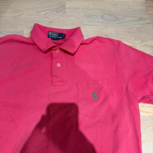 Rosa pikétröja från Polo Ralph Lauren - Snygg rosa pikétröja från Polo Ralph Lauren med klassisk krage, korta ärmar och knäppning framtill. Bröstficka med grön broderad logga. Perfekt för en chill och stilren look.
