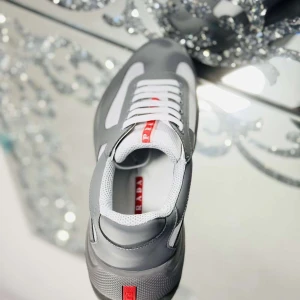 Prada cups grey - Snygga sneakers från Prada i grått och vitt med röda detaljer. Skorna har en modern design med meshpaneler, vita snören och en bekväm platt sula. Perfekta för dig som vill ha en clean och stilren look med exklusiv känsla.
