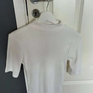 Vit ribbad polotröja med kort ärm - Säljer en stilren vit ribbad polotröja med korta ärmar. Tröjan har en högre hals och är gjord i ett stretchigt material som sitter snyggt på kroppen. Perfekt att matcha med jeans eller kjol för en clean look.