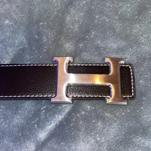 Svart H Bälte med H Spänne  - Säljer ett svart skärp med ikoniskt H-spänne i silverfärgad metall. Bältet är gjort i äkta skinn med vit kontrastsöm längs kanterna. Perfekt accessoar för att lyfta din outfit med en lyxig touch.