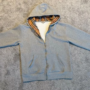  Burberry hoodie  - Snygg grå hoodie från Burberry med dragkedja och klassiskt rutigt foder i huvan. Jackan har broderad logga på bröstet, fickor fram och snörning i huvan. Perfekt för dig som gillar streetstyle och vill ha något som sticker ut.