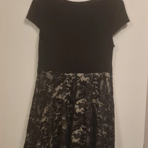 Svart spetsklänning från Bodyflirt M - Supersnygg svart kortklänning från Bodyflirt med blommiga spetsdetaljer och korta ärmar. Kjolen har svart spets över beige tyg och midjan är dekorerad med stora blommor. Perfekt för dig som vill sticka ut med en elegant look.