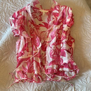 Blommig omlottblus från H&M - Supersöt blommig omlottblus från H&M i vitt och rosa med volangdetaljer och knyt framtill. Blusen är croppad och har korta ärmar samt v-ringning.  Den kommer inte till användning ❤️passar dig som är s också
