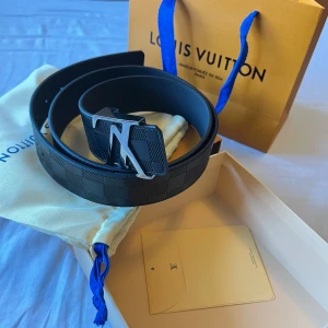 Louis Vuitton Bälte - Säljer ett svart bälte från Louis Vuitton med ikoniskt LV-spänne i svart metall. Bältet har diskret rutmönster och levereras i original ask, dustbag och påse med blå detaljer. Perfekt accessoar för att lyfta vilken outfit som helst. Använd fåtal gånger. Märks inte att den är använd. 110 cm. Köpt från Stockx