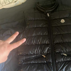 Moncler gui  - Säljer en svart, glansig dunväst från Moncler med hög krage och dragkedja framtill. Västen har två snedställda fickor med dragkedja och klassisk Moncler-logga på bröstet. Perfekt för lager-på-lager och snygg till streetwear. Kommer med påsen samt triangel skick 9-10 storlek 6skulle säga den passar mer som xl