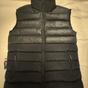 Svart dunväst från Philipp Plein M - Säljer en oanvänd snygg svart dunväst från Philipp Plein med quiltad design och dragkedja framtill. Västen har två fickor med dragkedja och coola logodetaljer på bröstet. Tillverkad i vattentät polyester och övre del i skinn.