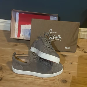 Grå mockasneakers från Christian Louboutin - Snygga grå sneakers från Christian Louboutin i mjuk mocka med klassisk vit sula och den ikoniska röda undersidan. Skorna har rund tå, snörning och stilren design som passar perfekt till streetwear eller en mer dressad look.