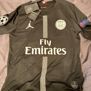 PSG Mbappé tröja - Svart PSG fotbollströja med Mbappé 7 på ryggen, tillverkad i samarbete med Jordan. Tröjan har vita detaljer, Fly Emirates-tryck, Champions League-märke på ärmen och PSG-logga på bröstet. Materialet är lätt och andas, perfekt för fotboll eller streetwear. Säljer för att den inte kommer till andvändning