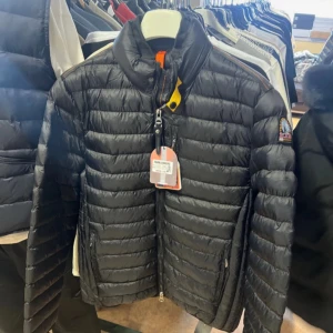 Svart dunjacka från Parajumpers M - Svart dunjacka från Parajumpers i storlek M. Jackan har quiltad design, hög krage och dragkedja framtill. Klassisk patch på ärmen och cool gul detalj vid halsen. Perfekt för kalla dagar och riktigt snygg till streetwear-stilen.