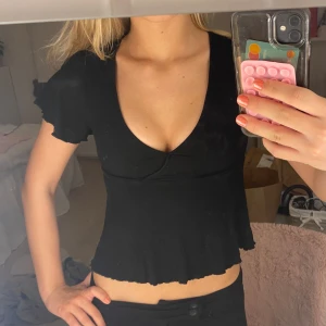 Svart ribbad u-ringad topp - Säljer en svart croppad topp med v-ringning och korta volangärmar. Toppen är ribbad och har en omlott-detalj vid bysten samt vågig kant nertill. Perfekt för dig som gillar en enkel men ändå trendig stil.
