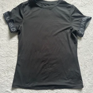 Svart t-shirt från Maya Delorez stl XS - Svart t-shirt från Maya Delorez med logga tryckt på ryggen och ärmarna. T-shirten har en klassisk passform, rund hals och är tillverkad i ett stretchigt syntetmaterial som känns sportigt och bekvämt. Perfekt för dig som gillar stilrena och sportiga plagg. Använd lite men inget märke/skav på den 