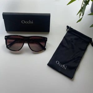 Bruna solglasögon från Occhi - Snygga solglasögon från Occhi med brunmelerat sköldpaddsmönster på bågarna och mörktonade glas. Modellen är klassisk med en lite större, fyrkantig form. Kommer med både hårt och mjukt fodral med Occhi-logga. Perfekta för soliga dagar. Aldrig använda! 
