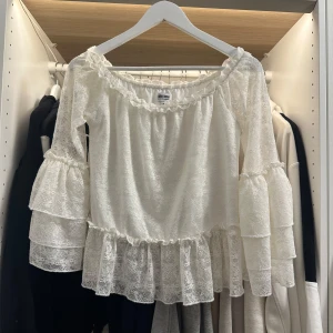 Vit spetsblus från Bubbleroom - Superfin vit blus från Bubbleroom med volangdetaljer och genombruten spets. Blusen har vida ärmar med flera lager volanger och kan bäras offshoulder. Perfekt för dig som gillar romantisk och feminin stil.