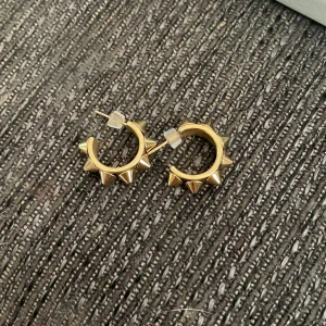Guldiga hoops med nitar - Coola guldiga örhängen i hoop-modell med spetsiga nitar runt hela ringen. Perfekta för dig som vill ha en edgy vibe och sticka ut lite extra. Säljs som ett par och har smidiga pluppar för att sitta säkert i örat.