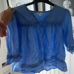 Blå blus med spets från Gina Tricot - Supersöt blå blus från Gina Tricot med v-ringning och fina spetsdetaljer längs bröstet och nederkanten. Blusen har trekvartsärm med rynkade muddar och är gjord i ett lätt och luftigt bomullstyg. Perfekt för dig som gillar en romantisk och chill stil.