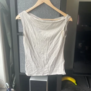 Beige ärmlös topp från Weekday - Säljer en stilren beige ärmlös topp från Weekday i mjuk bomull. Toppen har en asymmetrisk axel med knyt-detalj på ena sidan, vilket ger en cool och modern look. Perfekt att matcha med jeans eller kjol för en enkel men trendig outfit.