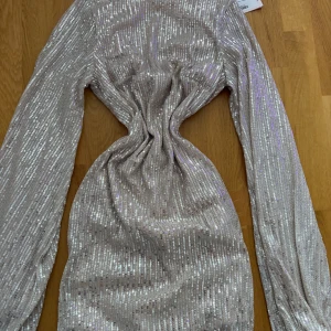 Glittrig kortklänning med paljetter - Supersnygg glittrig kortklänning i ljus champagnefärg med långa ärmar och paljetter över hela tyget. Klänningen har en tight passform och markerad midja som ger en cool siluett. Perfekt för dig som vill sticka ut och glänsa.