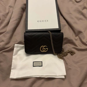 Svart Gucci Marmont axelväska i skinn - Säljer en svart Gucci Marmont axelväska i mjukt skinn med guldfärgad kedja och GG-logo framtill. Väskan har quiltad design med hjärtdetalj på baksidan och levereras med dustbag och originalbox. Perfekt storlek för det viktigaste och riktigt snygg till alla outfits.