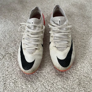 Nike Mercurial Air Zoom fotbollsskor - Säljer ett par Nike Mercurial Air Zoom fotbollsskor i vitt med svarta detaljer och röd häl. Skorna har snörning, låg profil och mönstrad sula för bästa grepp på planen. Ovandelen är i syntetmaterial och designen är riktigt sportig och modern.