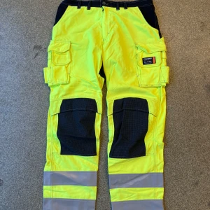 Tranemo arbetsbyxa, varsel och flamskyddade - Säljer ett par gula cargopants från Tranemo med svarta förstärkningar på knän och bak, samt stora fickor på sidorna. Byxorna har breda reflexband längst ner på benen och är tillverkade i slitstarkt bomullsmaterial. Perfekta för dig som vill sticka ut och synas.